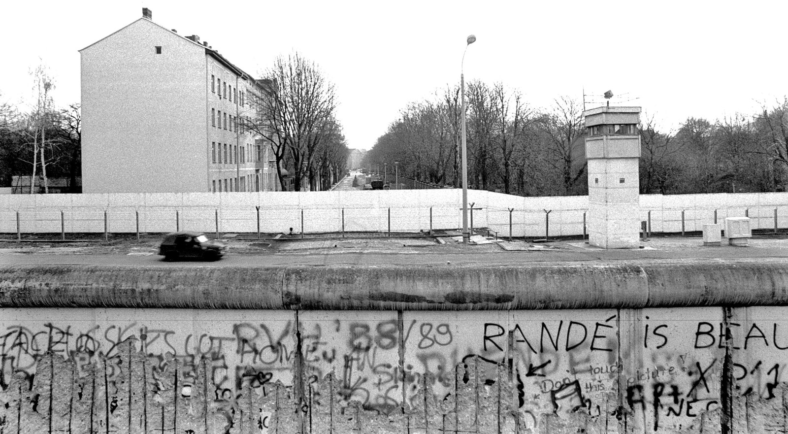 Black-and-white photo: in the foreground the rounded top of a graffiti-covered wall; beyond it a flat strip with a small car driving past, a square watchtower and an inner wall, with bare trees and apartment buildings behind.